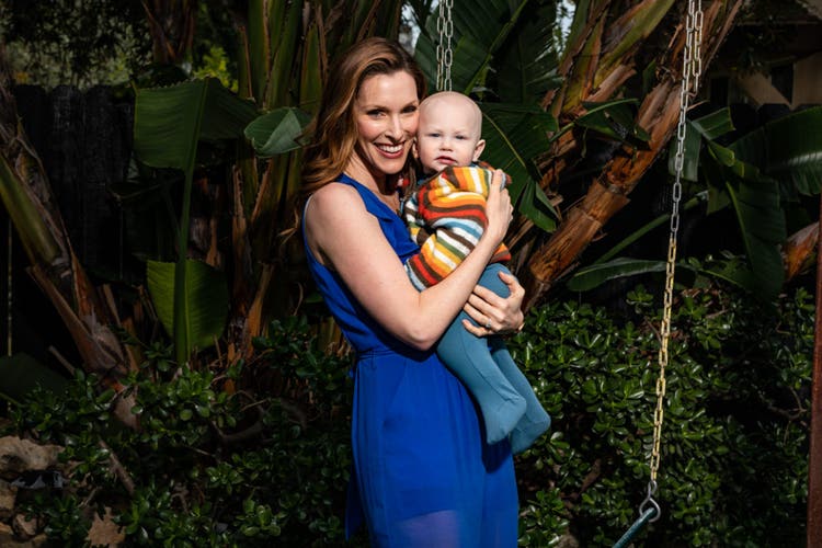 Emily Fletcher wears a blue short-sleeve dress and poses holding her infant child