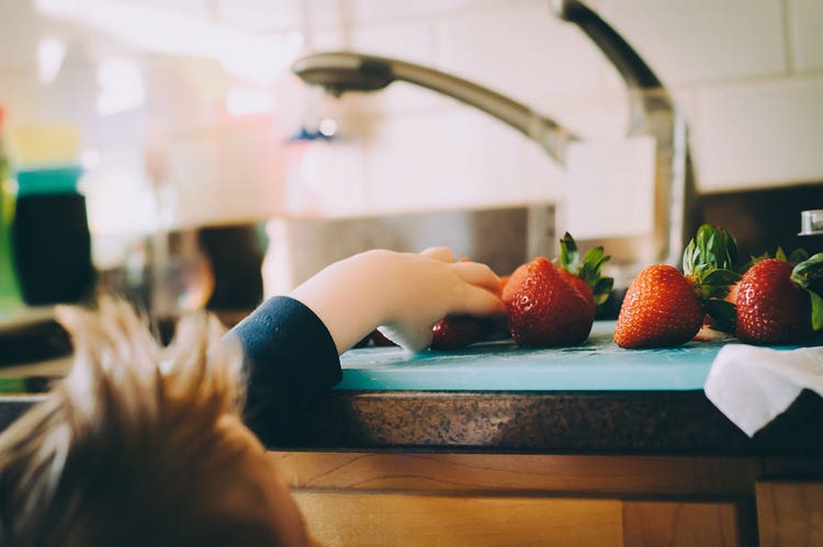 A small child reaches for a strawberry on a cutting board on a kitchen counter