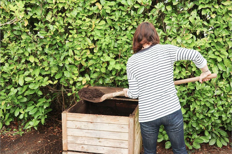 A person wearing jeans and a striped shirt shovels compost out of a wooden compost box