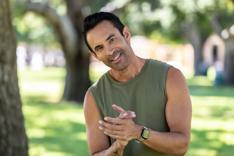 Jorge Cruise stands outside in a sunny park with hands clasped. Earthy green tank top