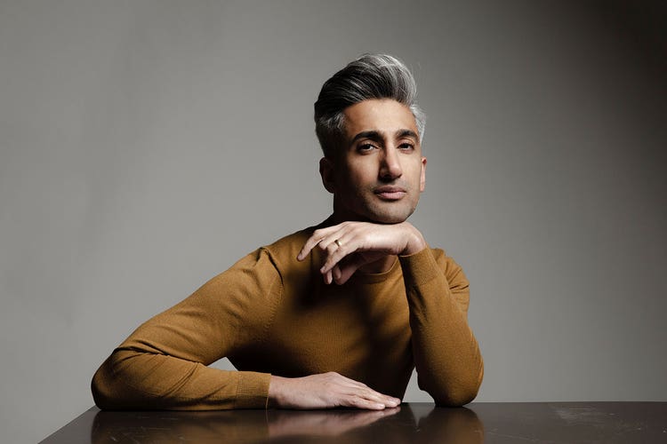 Tan France wearing a camel turtleneck sitting a glossy table, posed with a hand underneath his chin