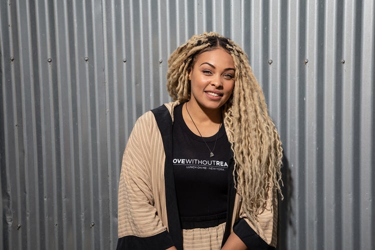Larayia Gaston stands against a corrugated metal backdrop and smiles. Her hair is twisted with long blonde extensions and she wears a tan striped jacket and cross necklace and tank that says: Love Without Reason.