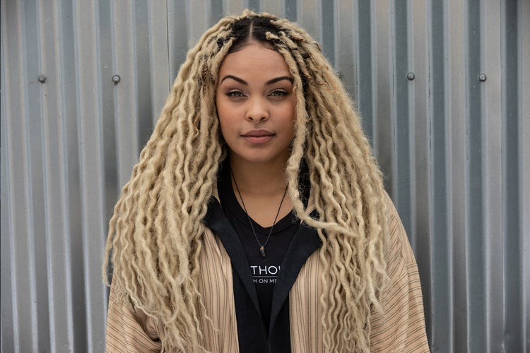 Larayia Gaston stands against a corrugated metal backdrop. Her hair is twisted with long blonde extensions and she wears a tan striped jacket and cross necklace.