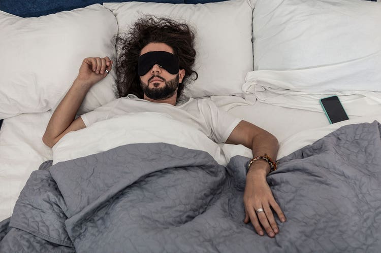 A person sleeps while wearing a black eye mask, underneath grey bedcovers in a brightly lit room