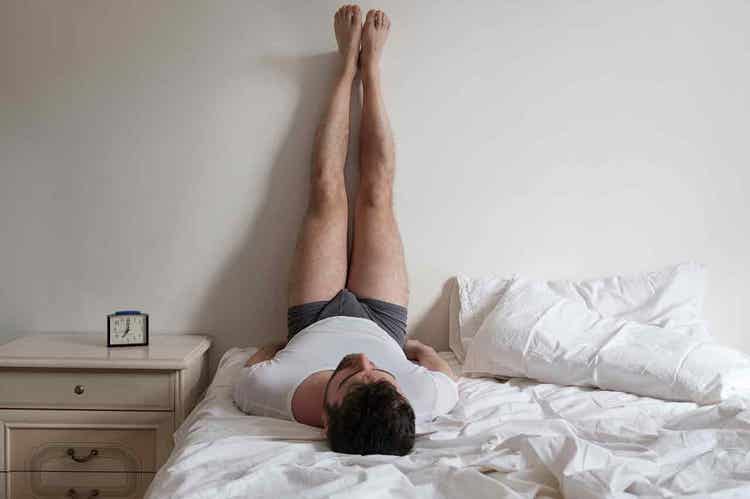 A person lays down on their bed with legs resting on the adjacent wall
