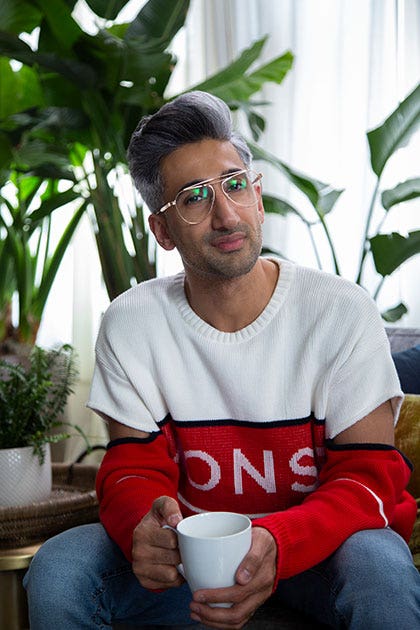 Tan sits on a couch holding a mug while wearing aviator glasses and a chunky knit sweater