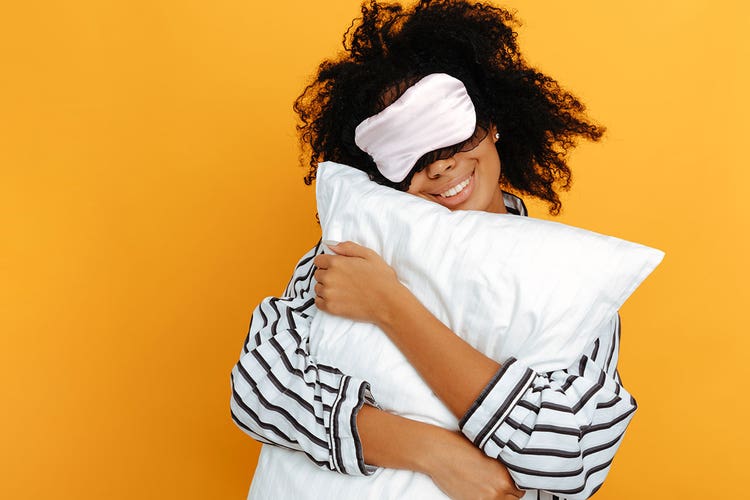 Person wearing striped black and white pajamas wears a matching eye mask and holds a white pillow against an orange backdrop