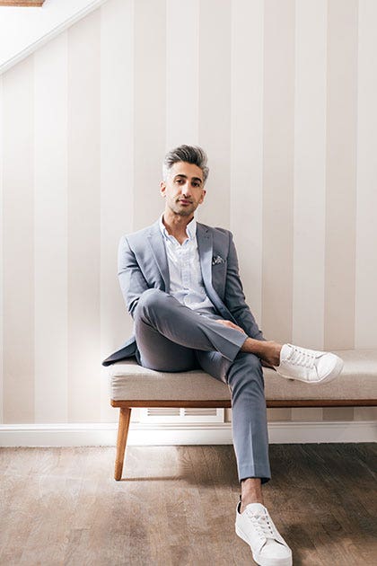 Tan France wearing a grey suit and pristine white shoes sitting on an ottoman